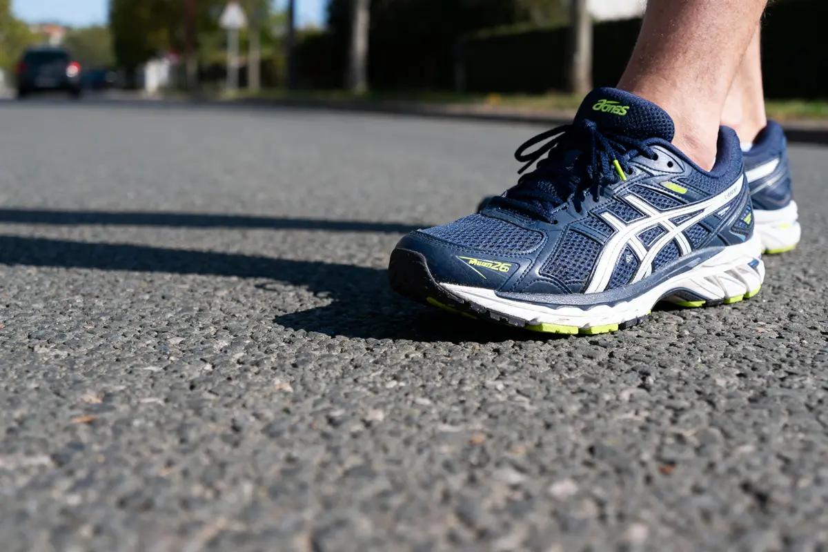 Un pied en baskets bleues Asics marche sur une route ensoleillée.