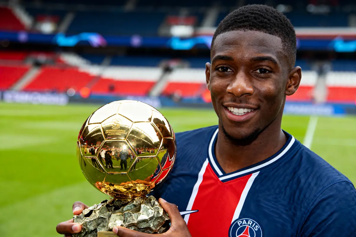 Joueur de football souriant, en tenue PSG, tenant un trophée doré sur un terrain de football.
