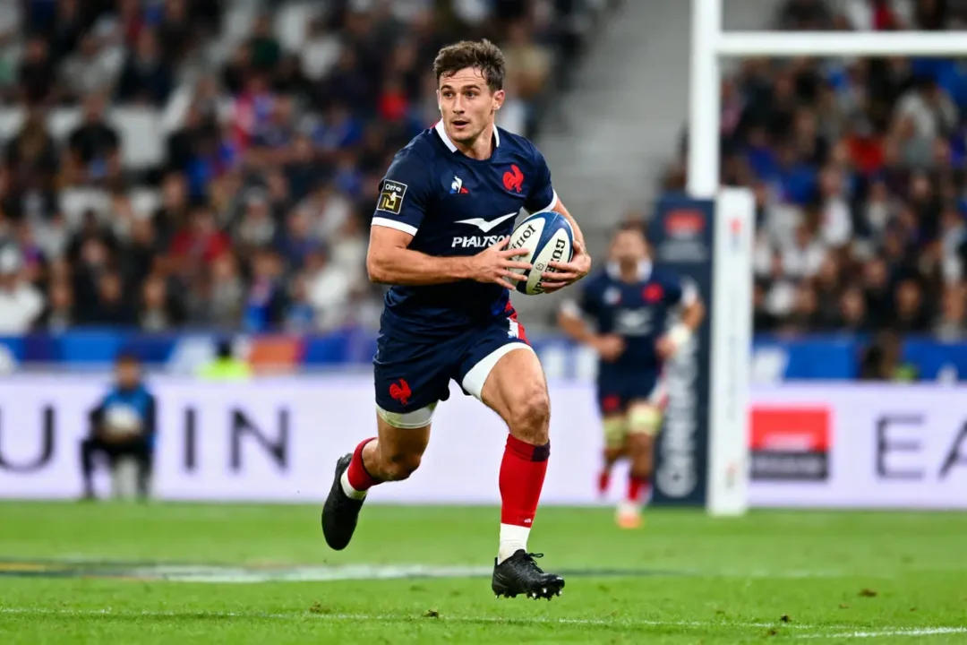 Joueur de rugby en action sur le terrain, tenant le ballon dans un stade rempli de spectateurs.