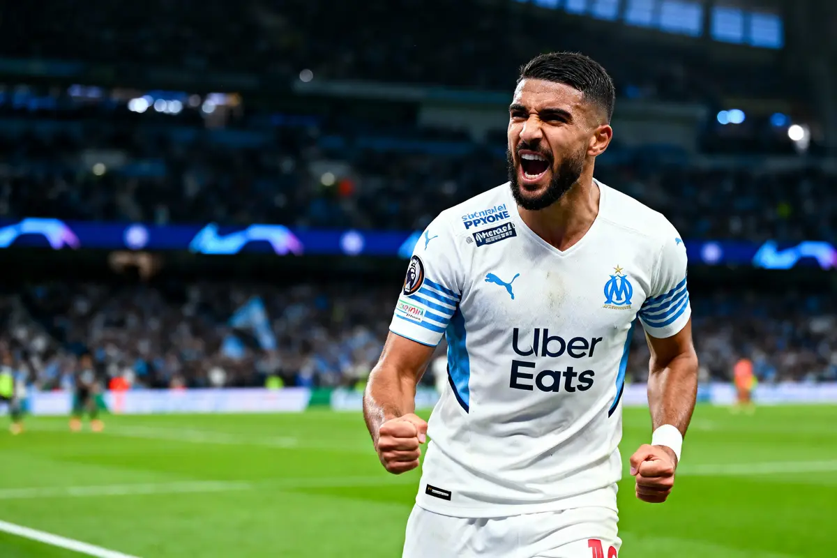 Joueur de football en maillot blanc célèbre un but sur le terrain, public en arrière-plan flou.