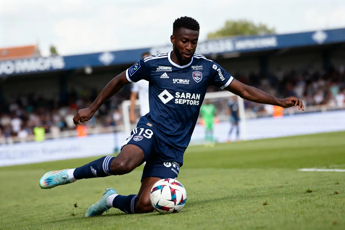Joueur de football en action, portant un maillot bleu, en train de tacler sur un terrain de football.