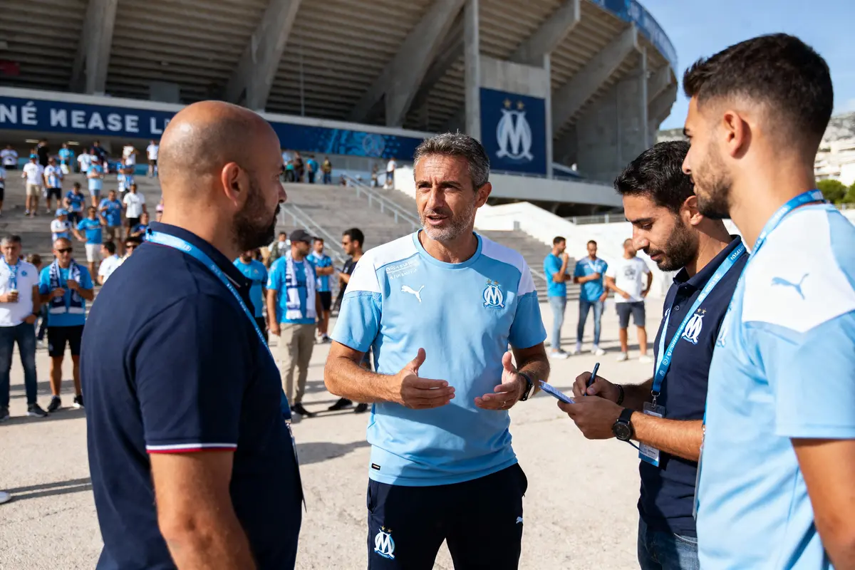Quatre hommes discutent devant un stade, portant des maillots de l'Olympique de Marseille.