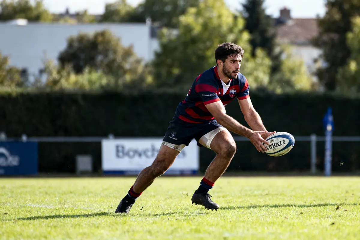 Joueur de rugby en action sur le terrain, tenant un ballon, en plein match, avec des arbres en arrière-plan.