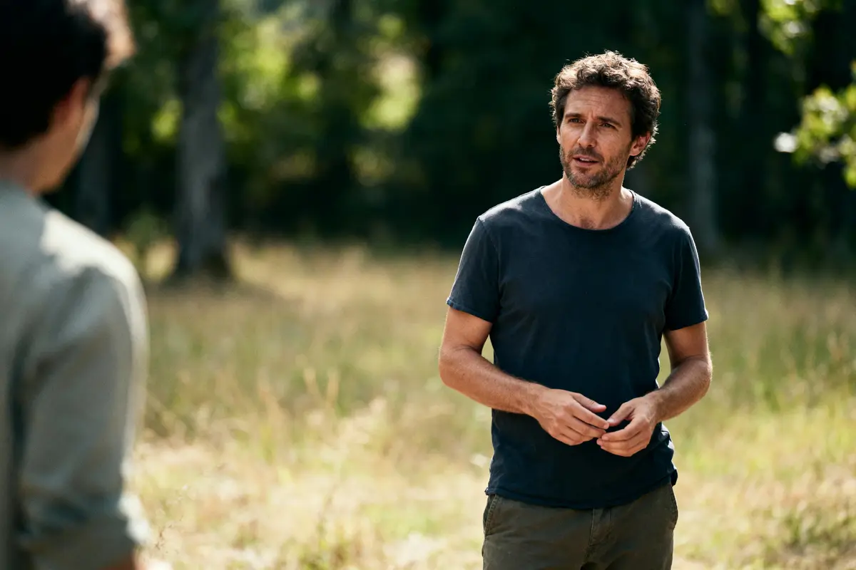 Homme debout, discutant en extérieur, t-shirt noir, environnement naturel en arrière-plan, journée ensoleillée.