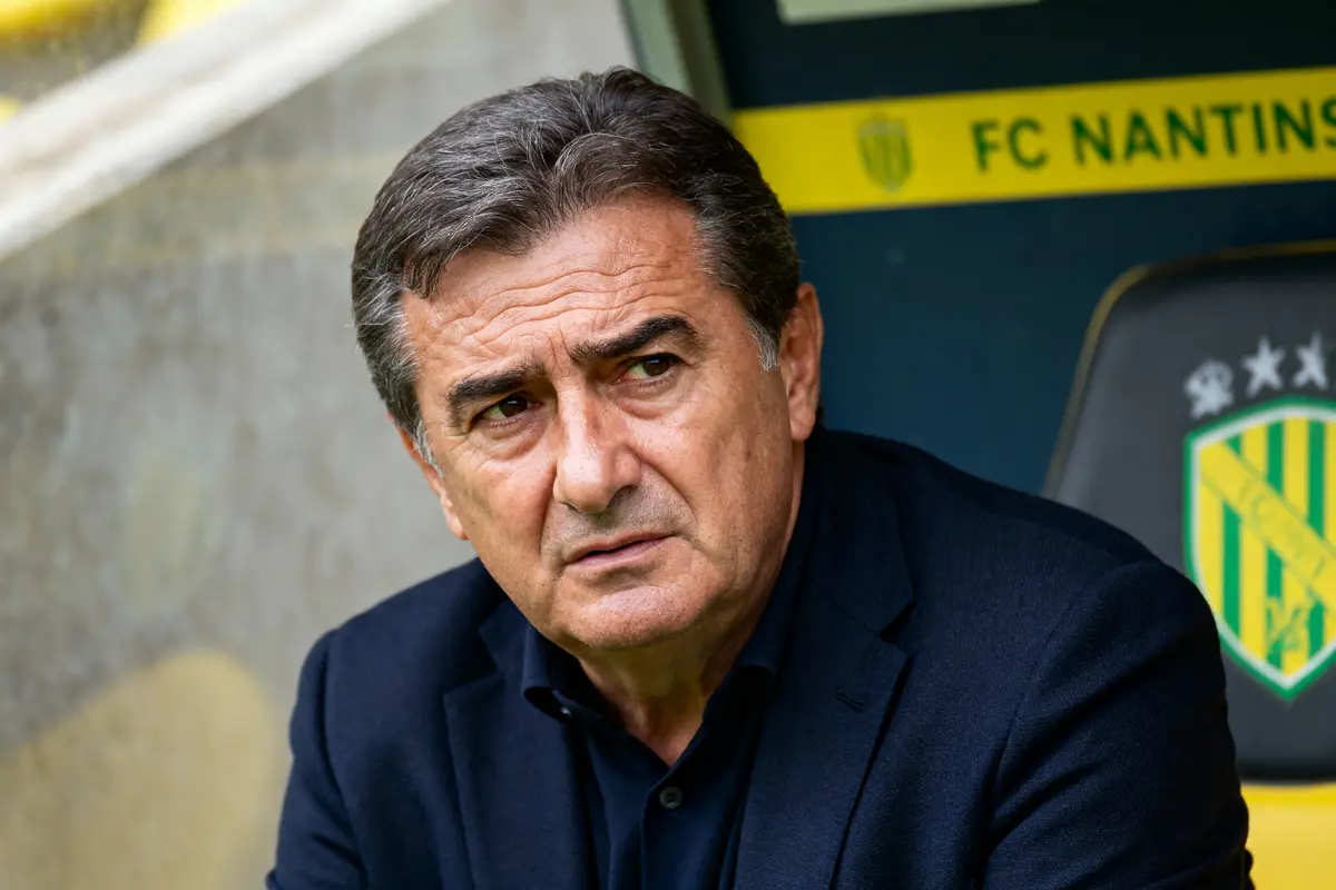 Homme pensif en costume sombre devant un panneau FC Nantes au stade.