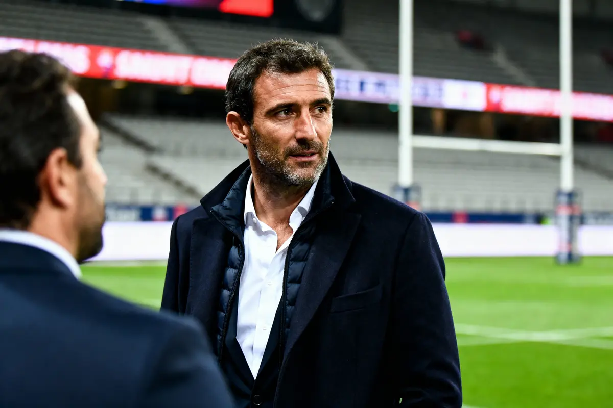 Un homme en costume se tient sur un terrain de rugby, stade en arrière-plan.