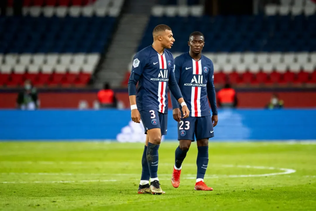 Deux footballeurs du PSG discutant sur le terrain, portant des maillots bleus avec bandes rouges et blanches.