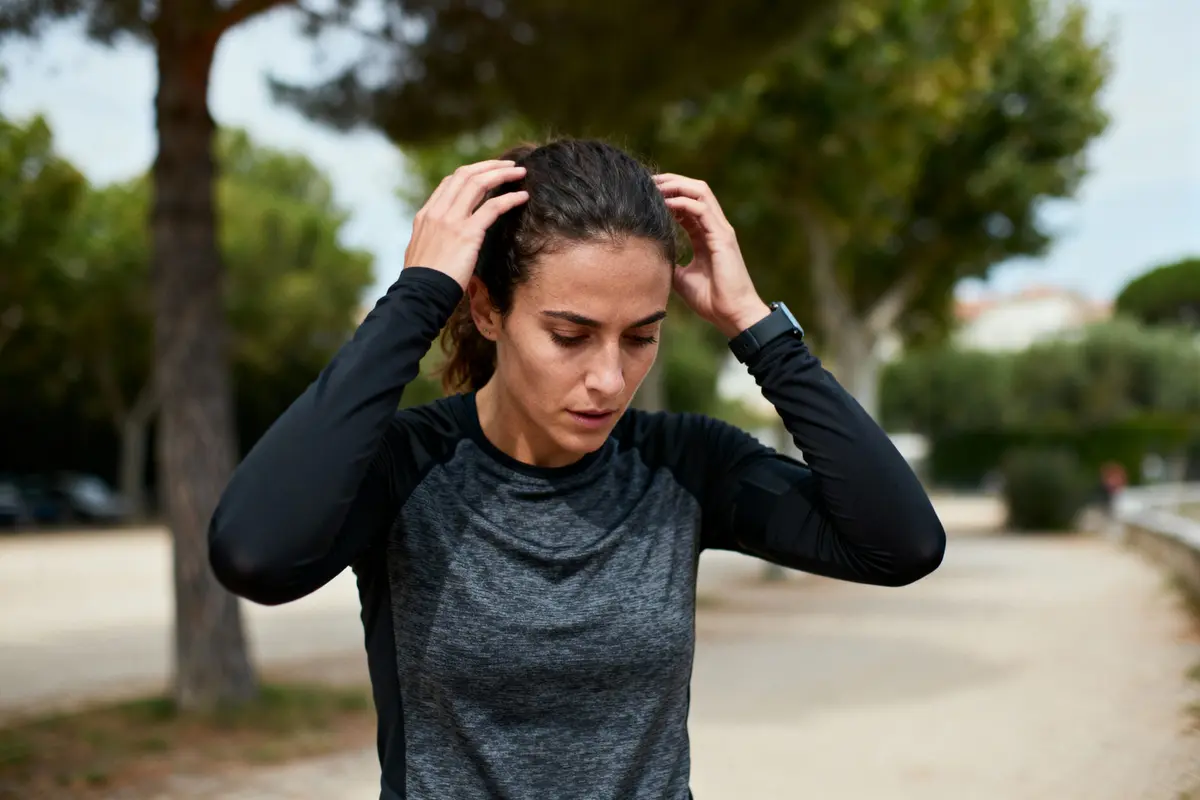 Femme en tenue de sport tenant ses cheveux, dans un parc arboré, l'air concentré.