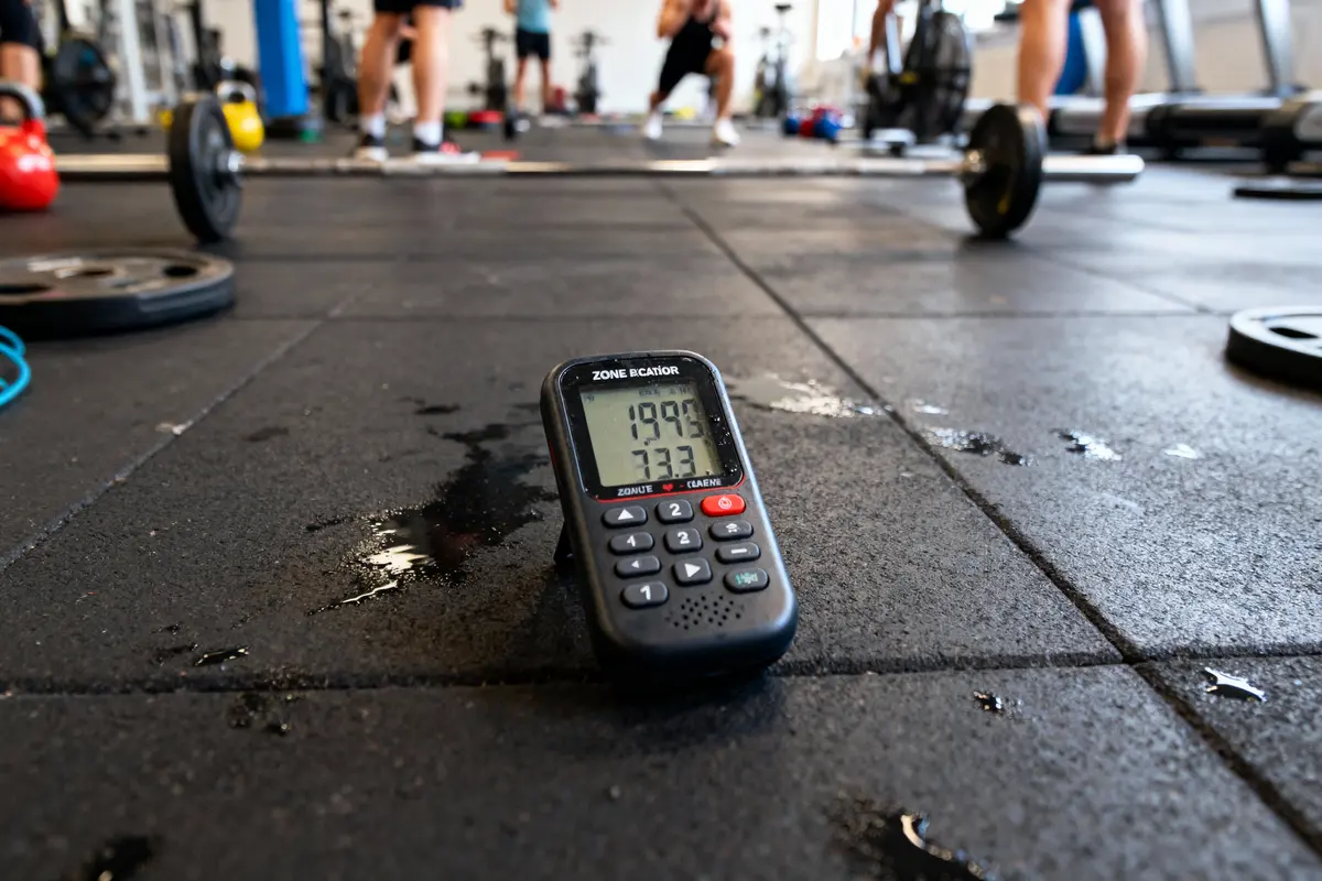 Chronomètre sur sol de gym avec haltères, personnes s’entraînant en arrière-plan. Ambiance de fitness dynamique.