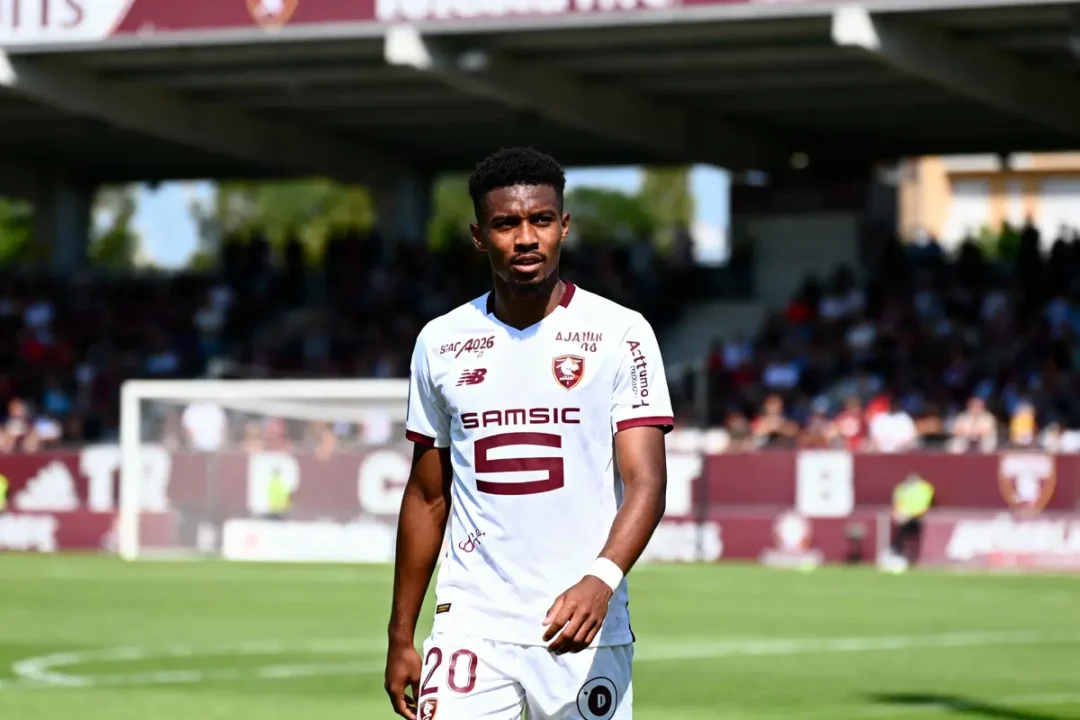 Joueur de football en maillot blanc sur le terrain, stade en arrière-plan avec spectateurs flous.