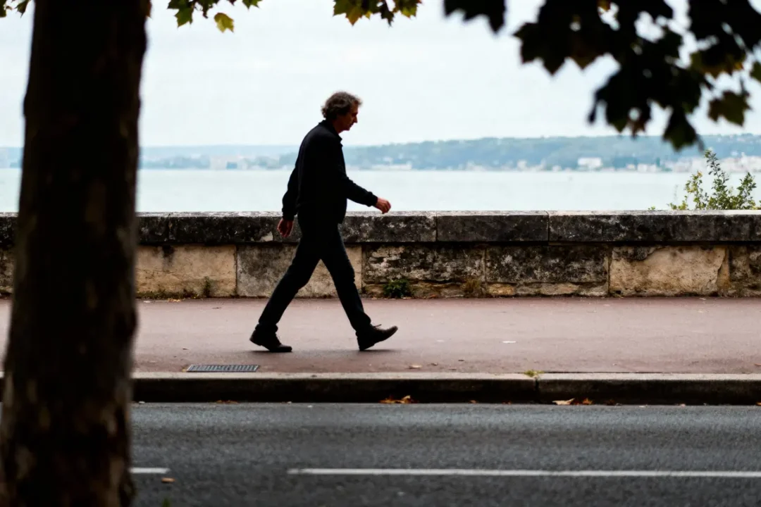 Homme marchant sur un trottoir près de la mer, derrière un mur de pierre, par une journée nuageuse.