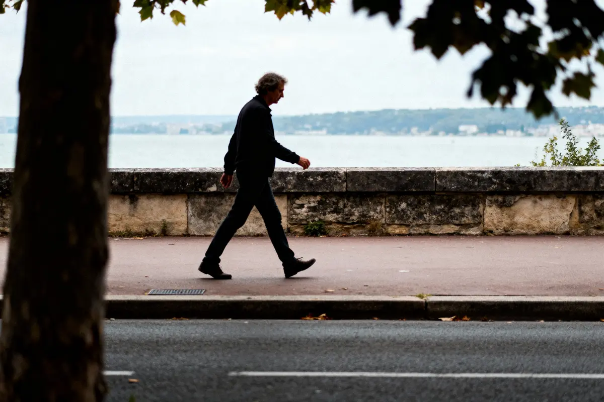 Homme marchant sur un trottoir près de la mer, derrière un mur de pierre, par une journée nuageuse.