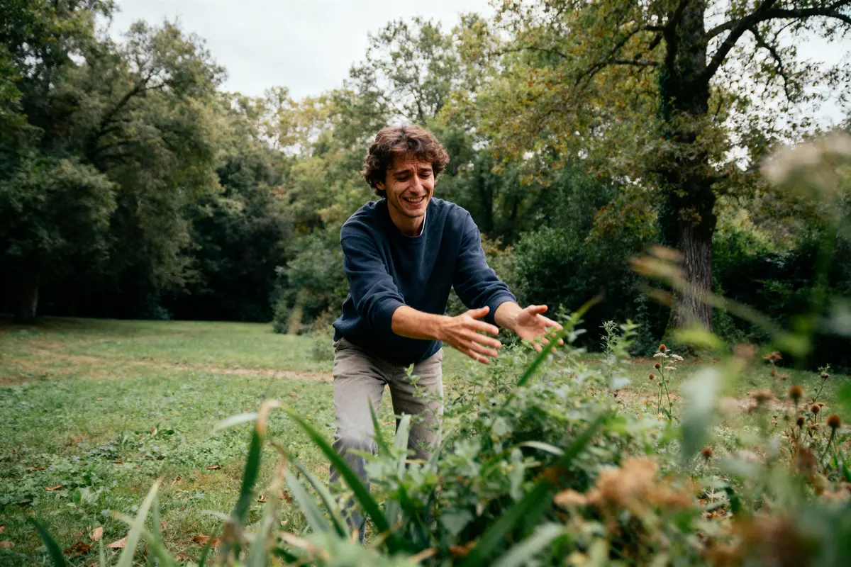 Homme souriant tend les bras dans la nature, entouré de verdure et d'arbres, en plein air.