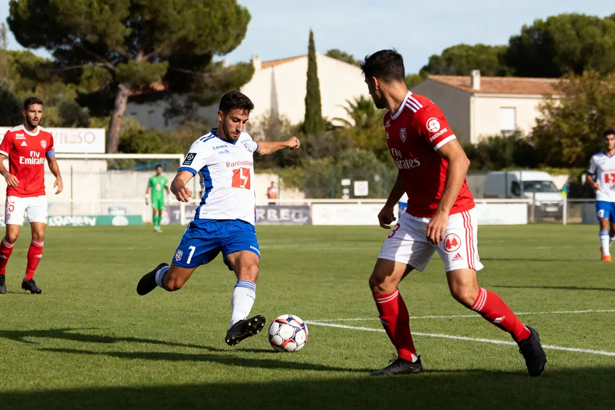 Deux joueurs de football disputent le ballon sur un terrain, entourés par des spectateurs et arbres.