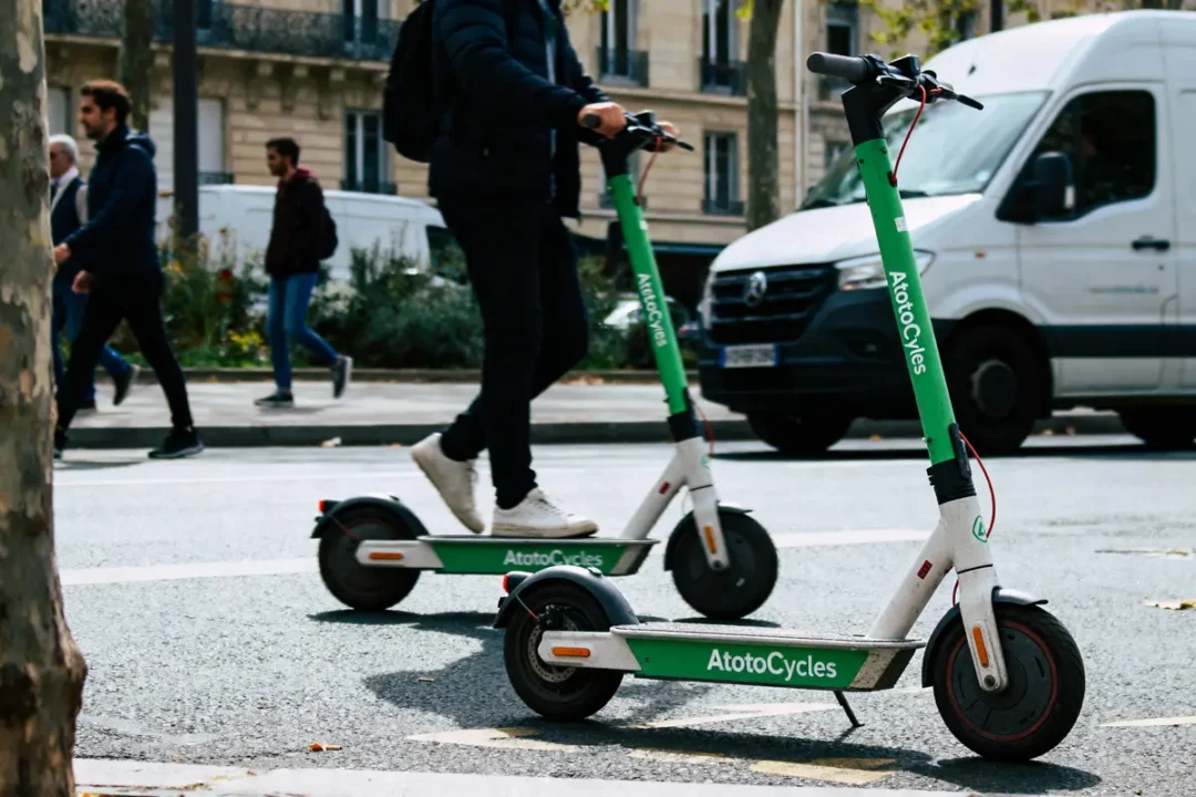 Deux personnes en trottinettes électriques vertes, rue urbaine avec piétons et camionnette blanche.