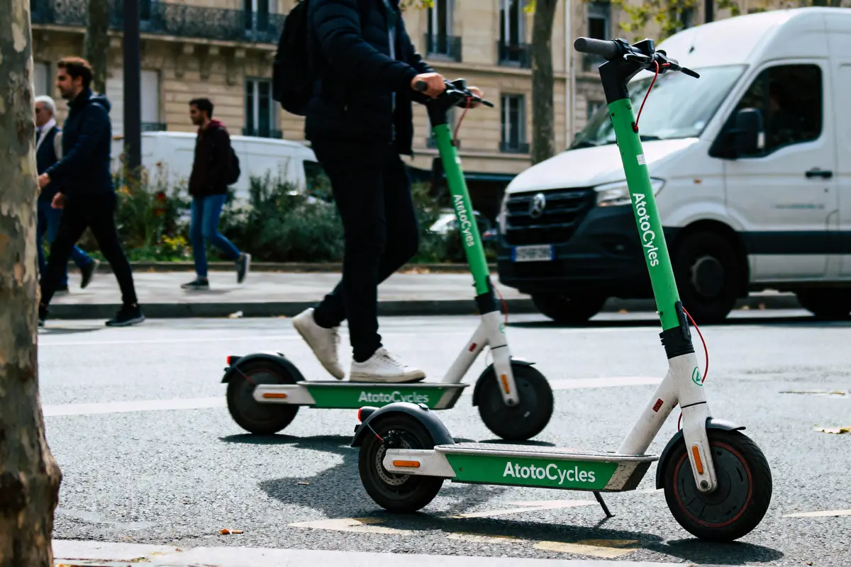 Deux personnes en trottinettes électriques vertes, rue urbaine avec piétons et camionnette blanche.