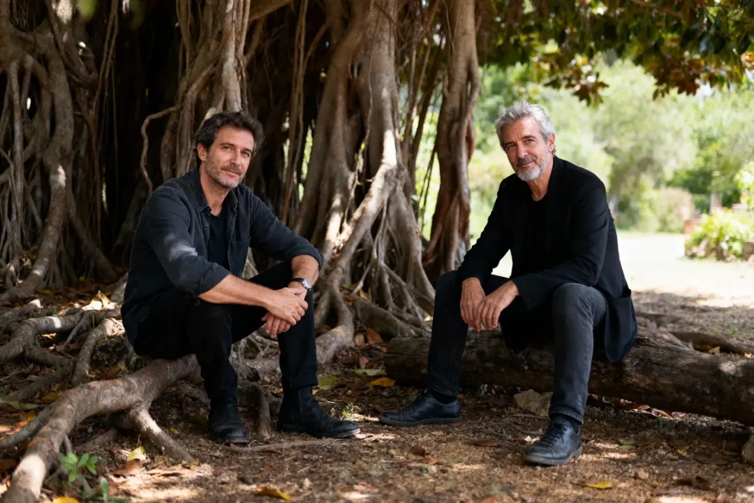 Deux hommes assis sous un grand arbre avec des racines apparentes, ambiance naturelle et détendue.