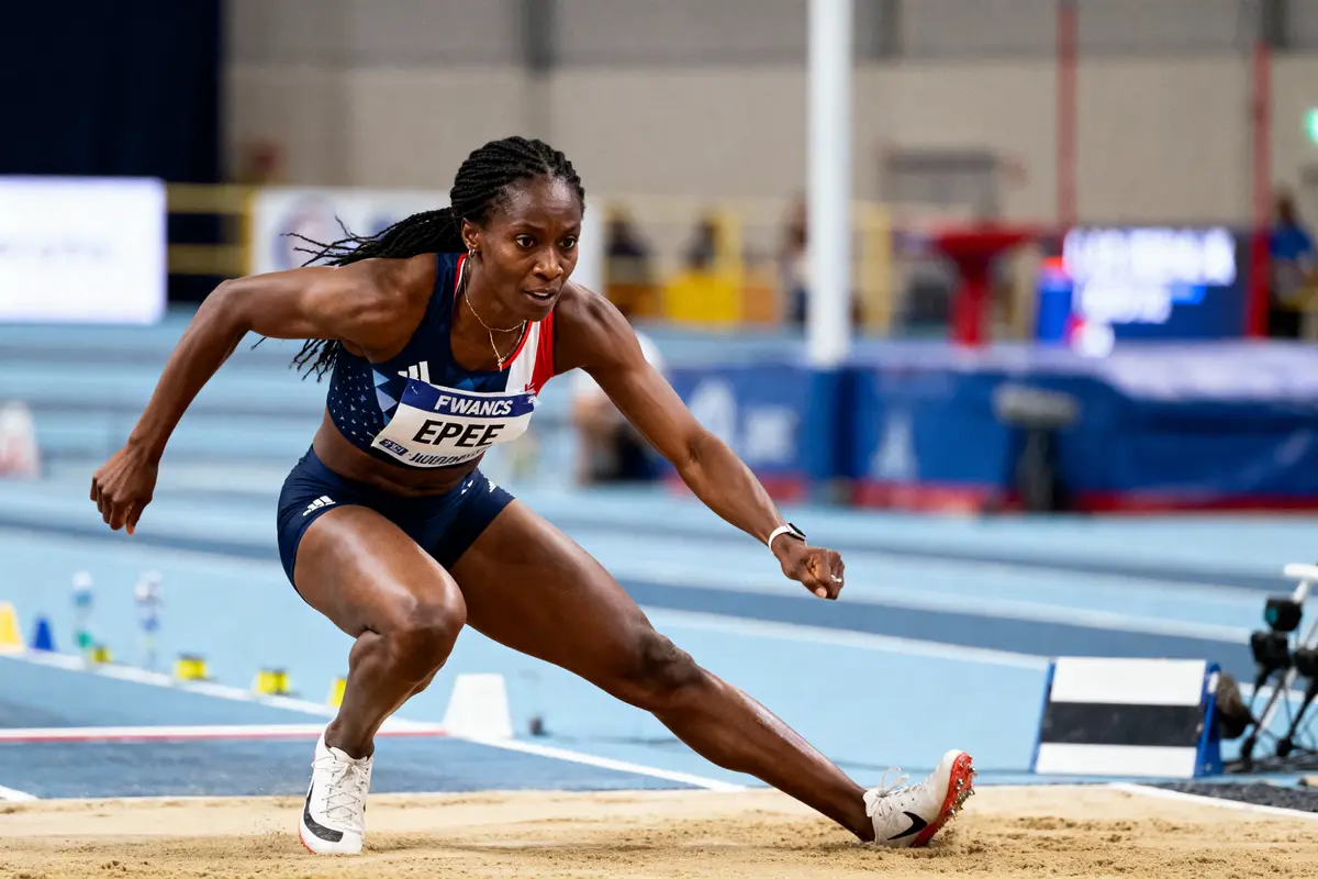 Athlète femelle atterrissant saut en longueur en salle, portant tenue bleue, concentrée, muscles tendus.