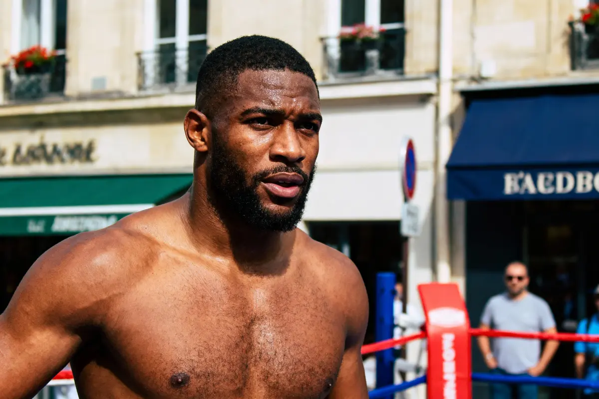 Homme torse nu dans un ring de boxe extérieur, bâtiments en arrière-plan, par une journée ensoleillée.