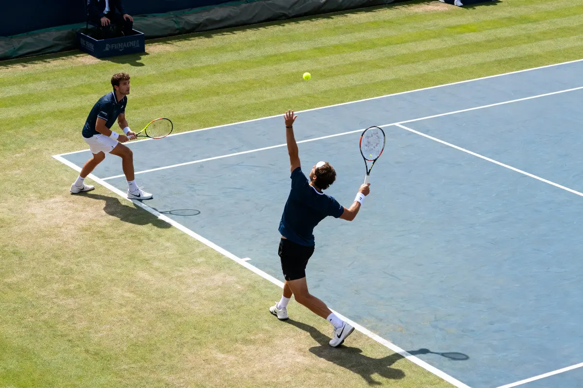 Deux joueurs de tennis en double sur gazon, l'un servant, l'autre prêt à recevoir.