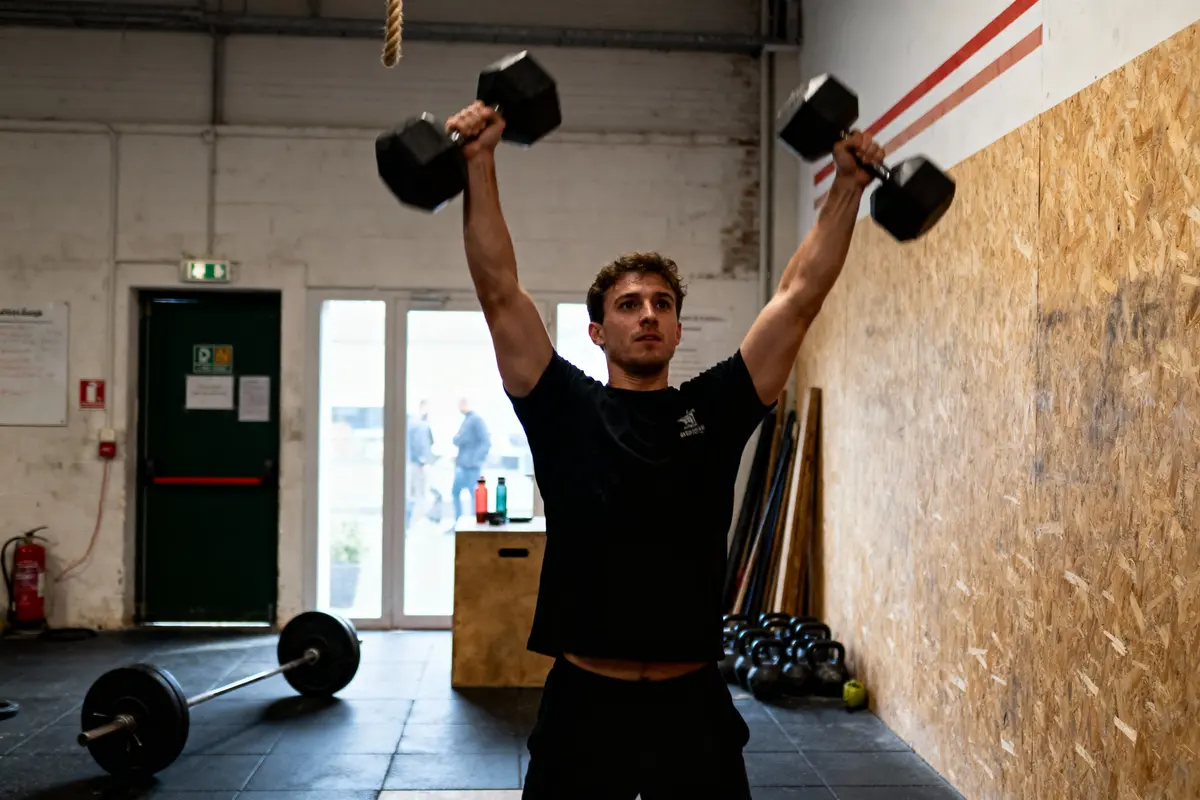 Homme levant des haltères dans une salle de musculation, ambiance sombre avec équipement de sport autour.
