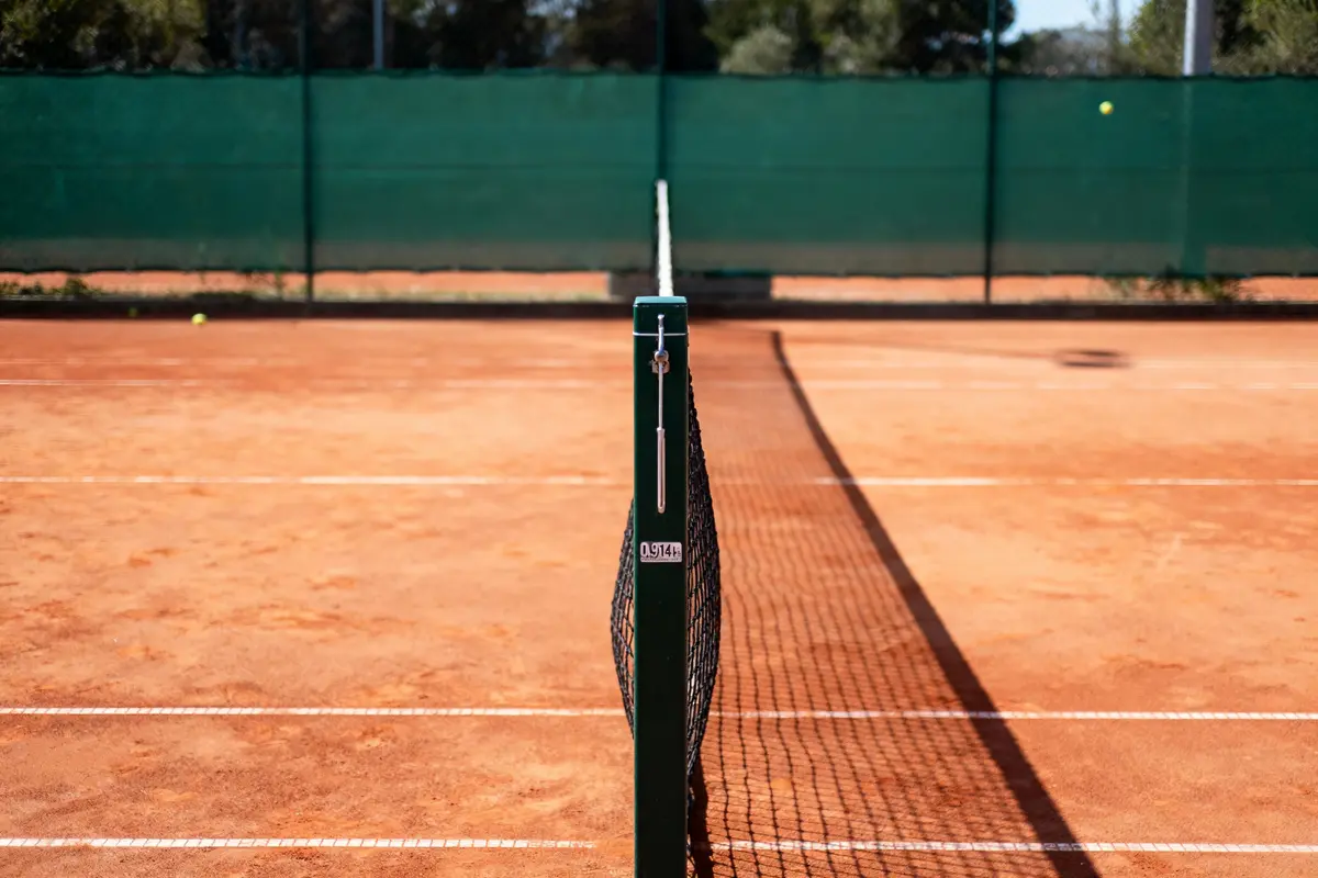 Court de tennis en terre battue, filet central, lignes blanches, ombre légère, clôture verte en arrière-plan.