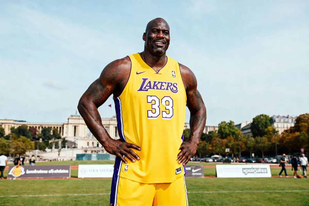 Un homme en maillot jaune des Lakers 33, debout sur un terrain avec bâtiments historiques en arrière-plan.