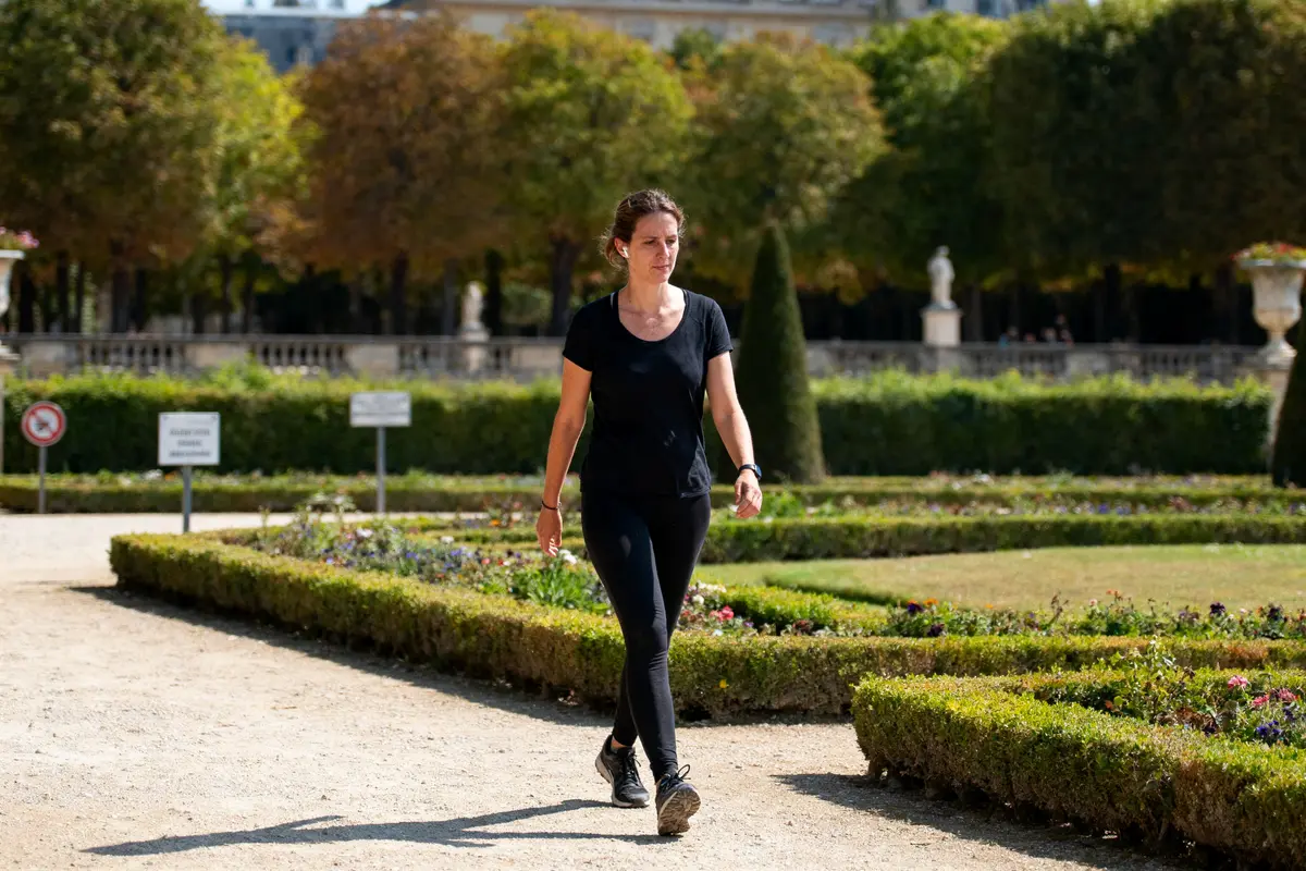 Femme marchant dans un parc verdoyant et ensoleillé, entourée de haies et de fleurs.