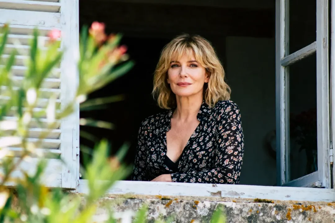 Femme souriante aux cheveux blonds, debout à une fenêtre, derrière des volets blancs et une plante verte.