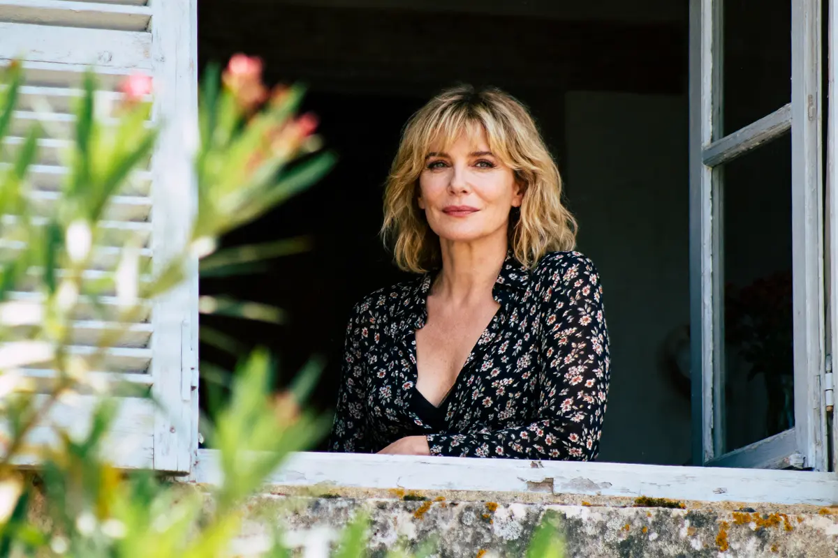Femme souriante aux cheveux blonds, debout à une fenêtre, derrière des volets blancs et une plante verte.