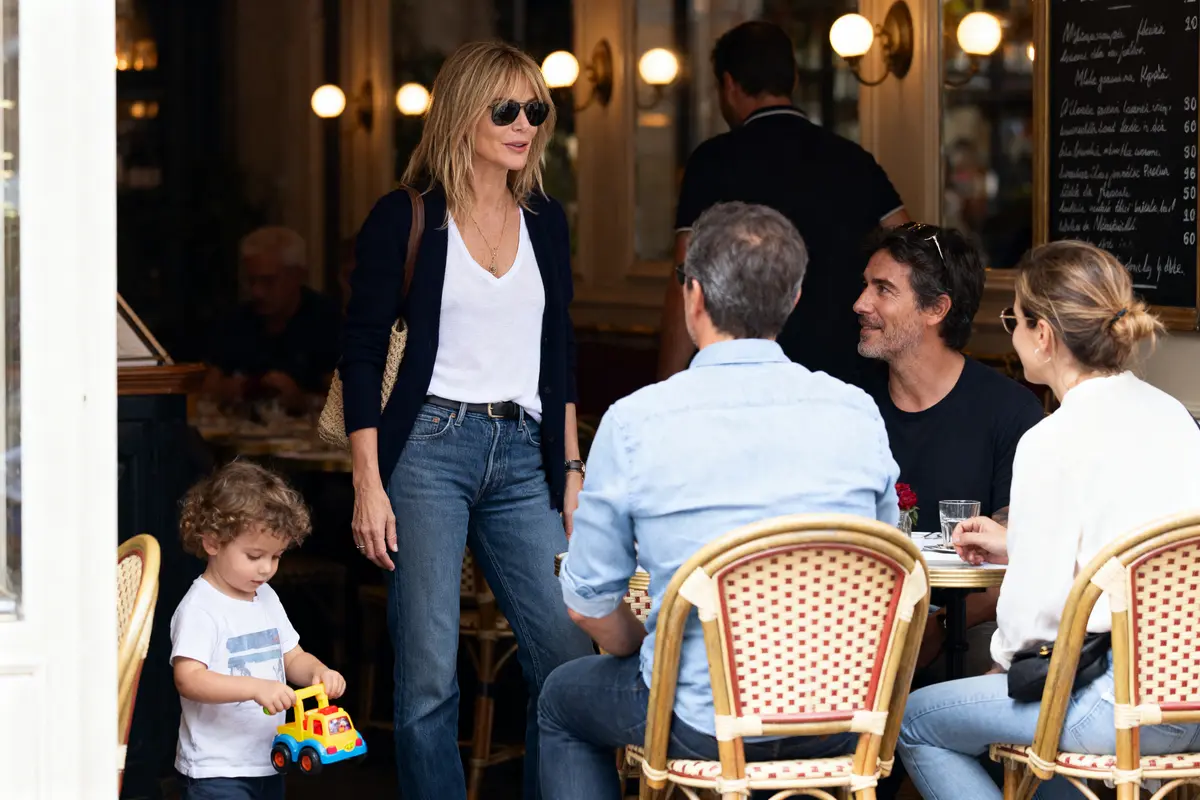 Une femme avec lunettes de soleil se tient debout près d'une table occupée, enfant jouant avec un jouet.