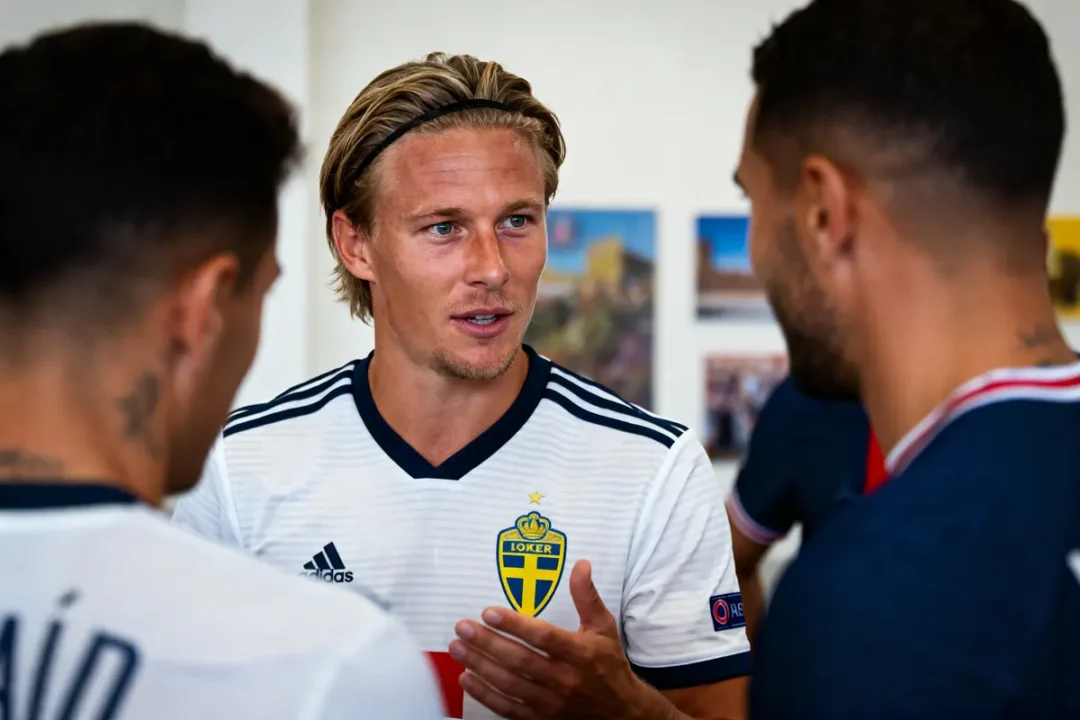 Trois hommes discutant, l'un portant un maillot blanc de l'équipe suédoise de football, en intérieur.