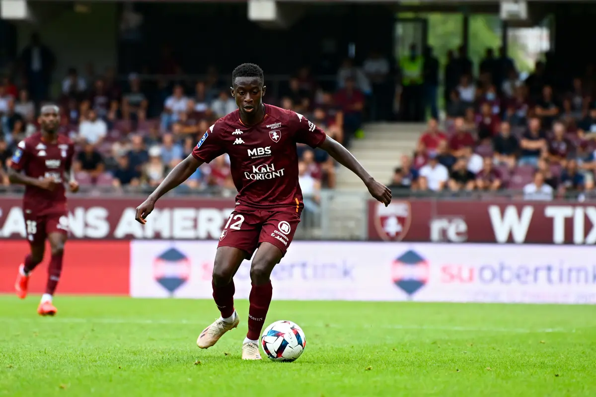 Jeune joueur de football en action sur le terrain, portant un maillot grenat, devant un public en tribunes.