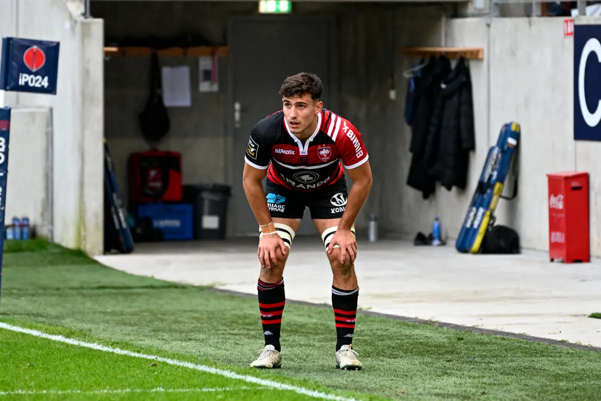 Joueur de rugby en maillot rouge et noir sur le terrain, mains sur les genoux, concentré.
