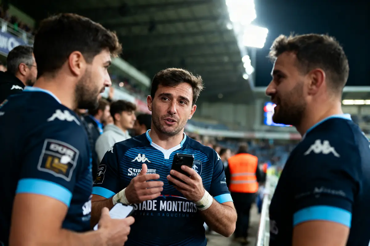 Trois joueurs de rugby discutent sur le terrain, portant des maillots bleus.