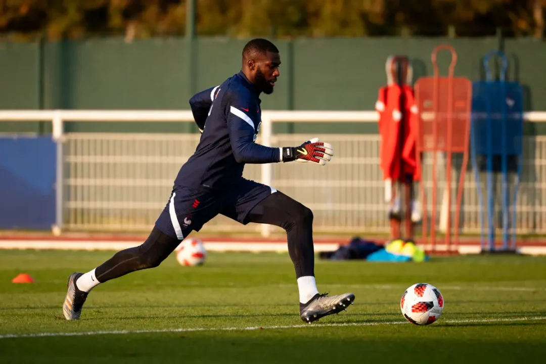 Footballeur en tenue d'entraînement, courant sur le terrain avec un ballon, équipements en arrière-plan.