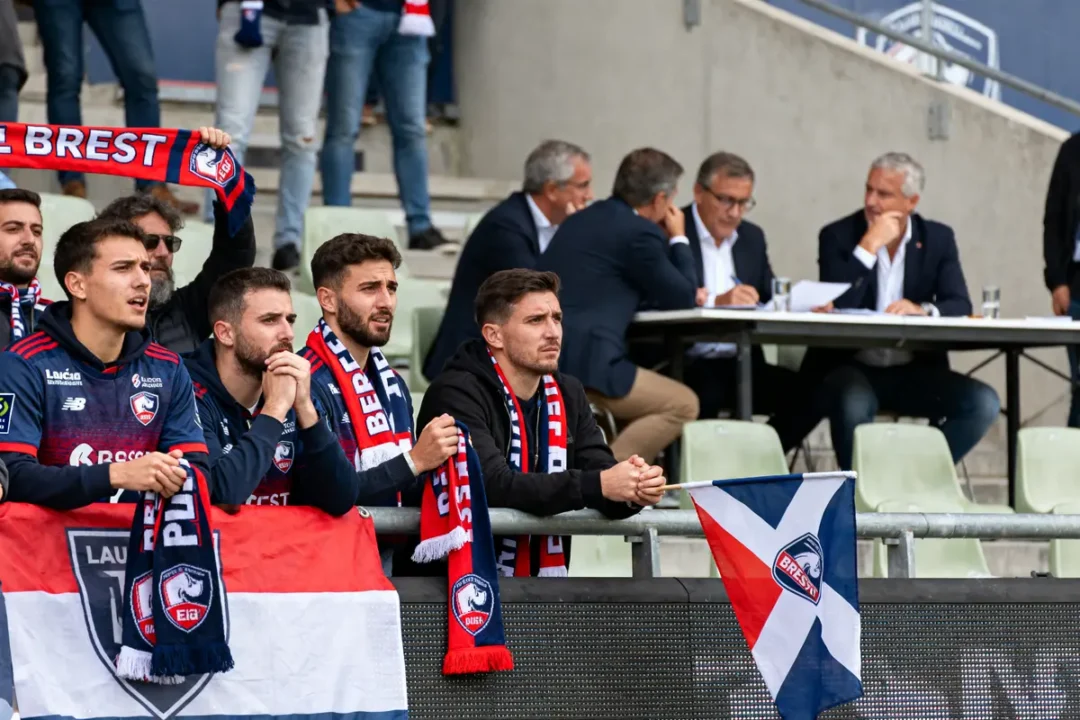 Supporters de football dans les tribunes avec écharpes et drapeaux de Brest, hommes en costume à l'arrière-plan.