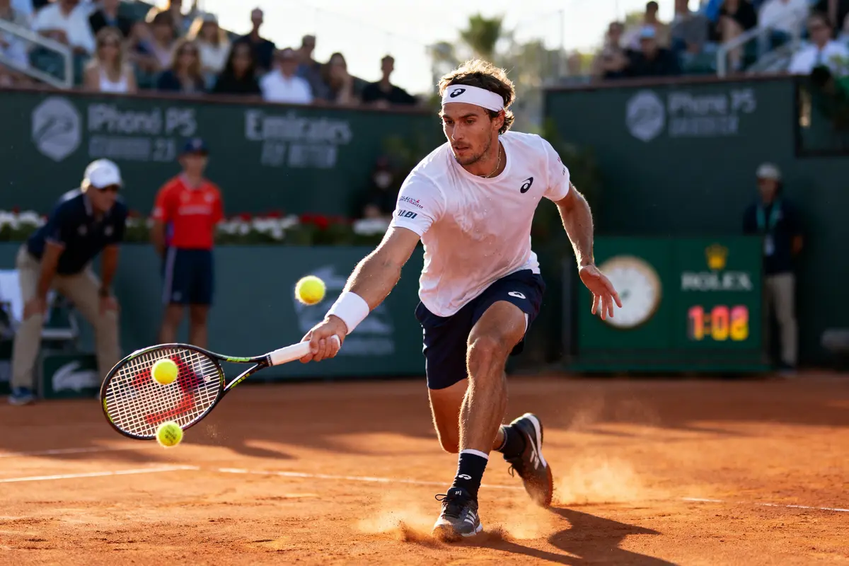 Joueur de tennis en action sur terre battue, exécutant un revers. Spectateurs et arbitres en arrière-plan.